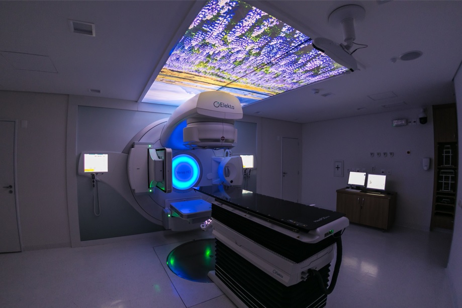 O Acelerador Linear, uma máquina branca com uma maca para fazer radioterapia; no teto, uma paisagem de campo para relaxar pacientes