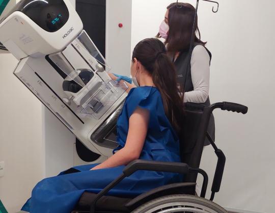 Moça branca sentada na cadeira de rodas com o mamógrafo abaixado